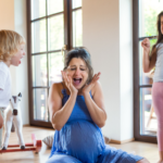 Here is the alt text in English: A pregnant mother wearing a blue dress sits on the floor with her hands on her face, looking overwhelmed and screaming. On the left, a young boy in a white shirt and beige shorts is shouting at her, while on the right, a young girl in a gray shirt and pink pants is throwing a tantrum with clenched fists and eyes shut tight. They are in a bright living room with large glass doors in the background.