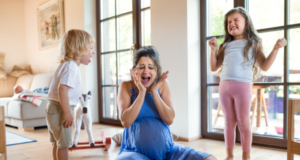 Here is the alt text in English: A pregnant mother wearing a blue dress sits on the floor with her hands on her face, looking overwhelmed and screaming. On the left, a young boy in a white shirt and beige shorts is shouting at her, while on the right, a young girl in a gray shirt and pink pants is throwing a tantrum with clenched fists and eyes shut tight. They are in a bright living room with large glass doors in the background.
