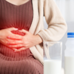 Medium shot of a woman sitting down, holding her stomach area with both hands. A red graphic glow over her abdomen symbolizes pain or discomfort. In the foreground, a glass of milk and a plastic bottle with a blue cap sit on a table.