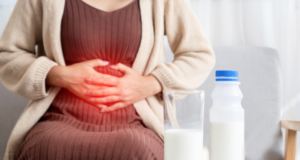 Medium shot of a woman sitting down, holding her stomach area with both hands. A red graphic glow over her abdomen symbolizes pain or discomfort. In the foreground, a glass of milk and a plastic bottle with a blue cap sit on a table.