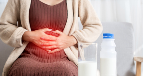 Medium shot of a woman sitting down, holding her stomach area with both hands. A red graphic glow over her abdomen symbolizes pain or discomfort. In the foreground, a glass of milk and a plastic bottle with a blue cap sit on a table.