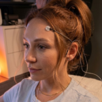 A high-resolution photo of a woman sitting in profile with hair pulled back in a high bun. She has a medical or research sensor electrode attached to her left temple with thin wires extending behind her ear. Warm light glows softly on her face from ambient lamps in the background, including a salt lamp on a bedside table or cabinet.