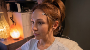 A high-resolution photo of a woman sitting in profile with hair pulled back in a high bun. She has a medical or research sensor electrode attached to her left temple with thin wires extending behind her ear. Warm light glows softly on her face from ambient lamps in the background, including a salt lamp on a bedside table or cabinet.